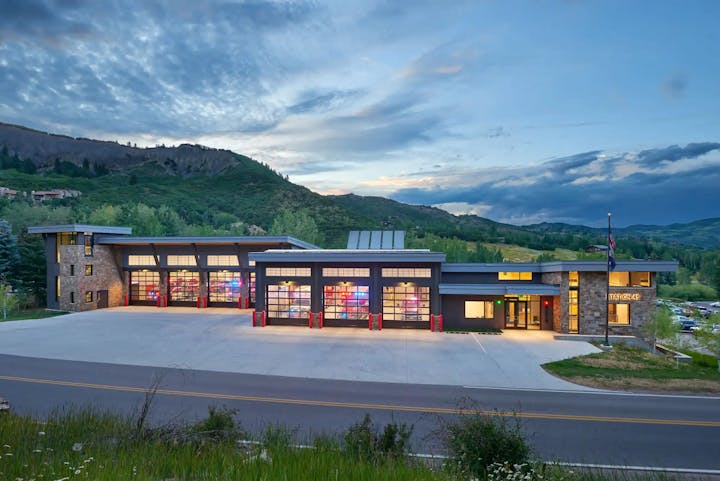 A modern fire station at dusk, with illuminated garage doors and a tranquil mountain backdrop.