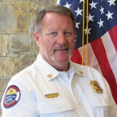 A man in a white fire department uniform with badges in front of the US flag.