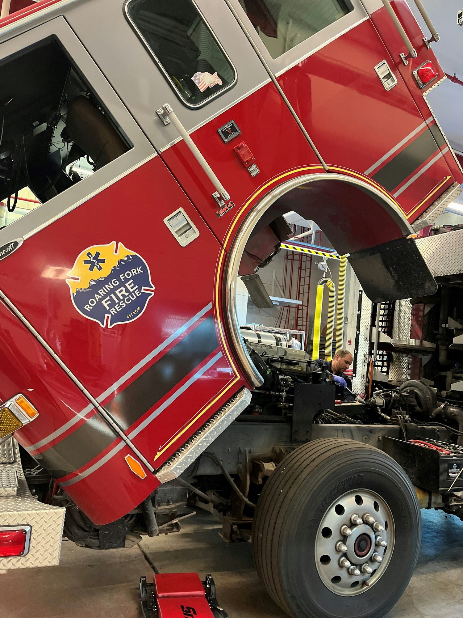 A fire truck in a garage with its engine exposed, and a mechanic working on the truck.