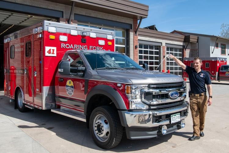 A firefighter standing next to a red fire department vehicle with "ROARING FORK" written on it, in front of a fire station.