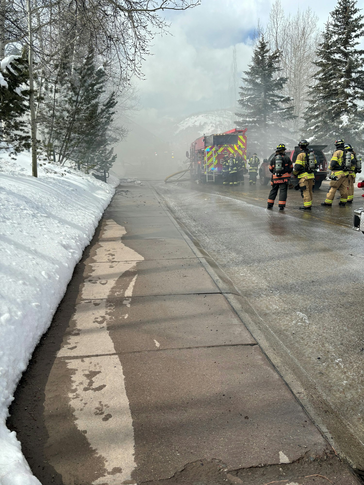 Firefighters near fire truck on snowy street with smoke in the background.