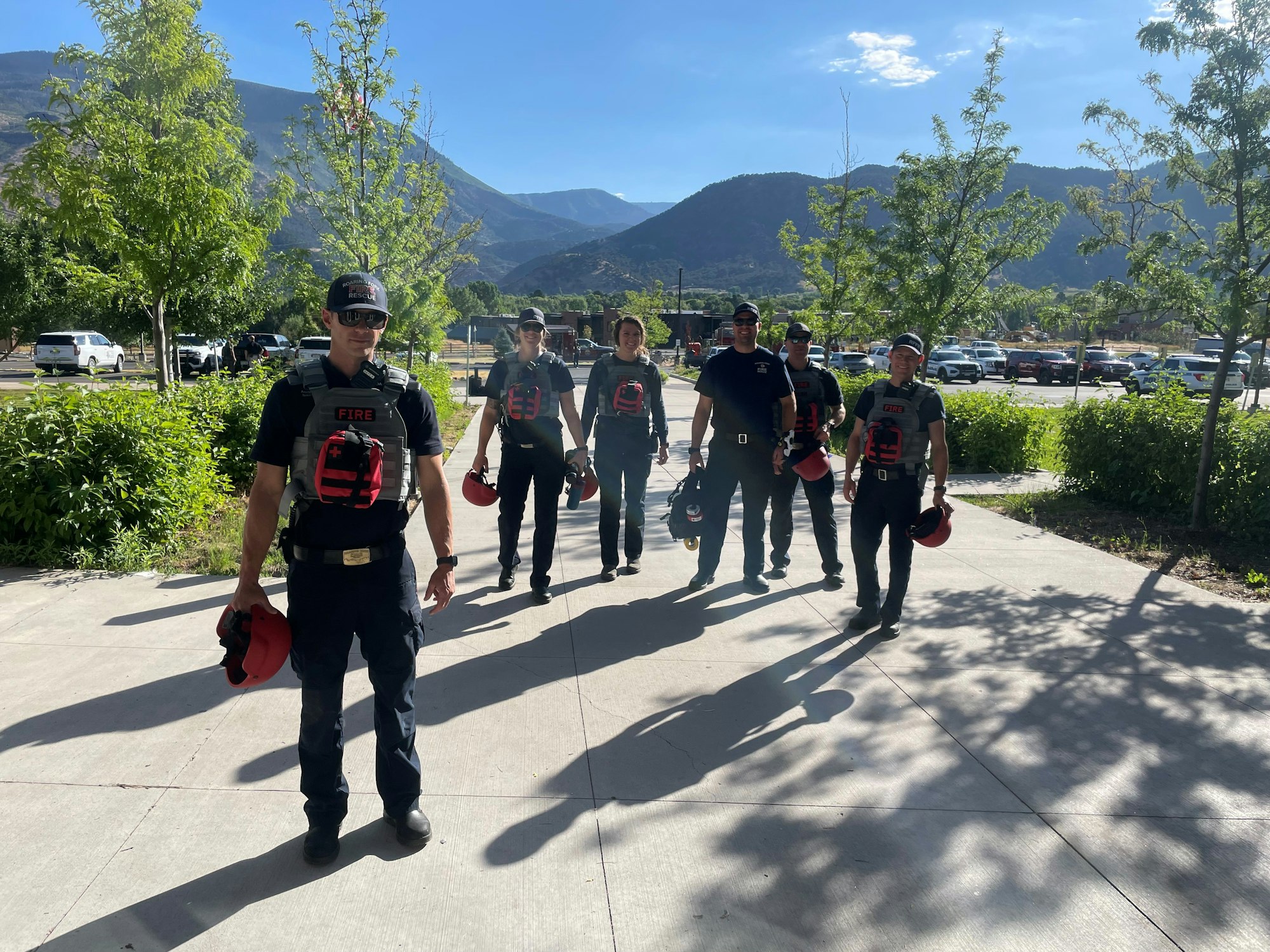 A group of firefighters walking outside, wearing uniforms and carrying helmets, with mountains in the background.