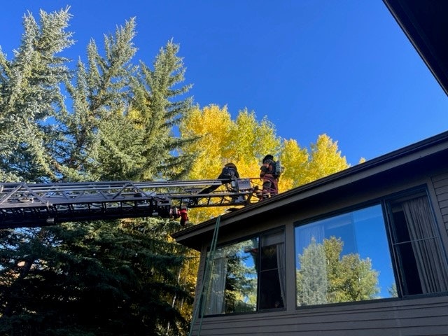 Firefighters on a ladder truck beside a house, with trees showing autumn colors in the background.