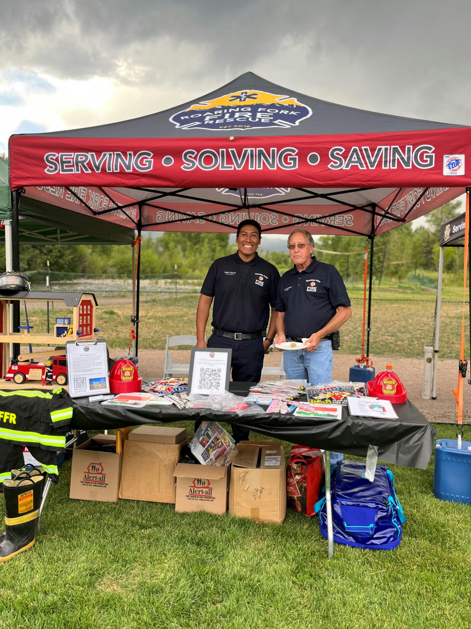 Two firefighters at an outreach booth with educational materials and fire safety items under a tent.