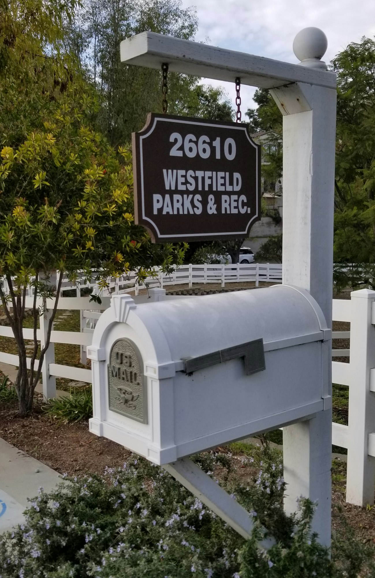 White mailbox mounted on a post with an overhead sign reading "26610 WESTFIELD PARKS & REC." The mailbox is situated in front of a white fence and surrounded by greenery.