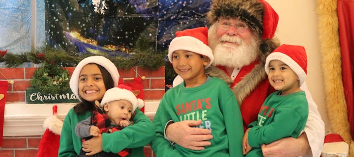 A group of children with Santa hats smiling with a person dressed as Santa Claus against a festive backdrop.