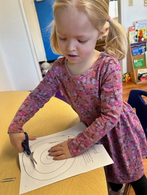 A child in a patterned dress is cutting paper with scissors on a table.