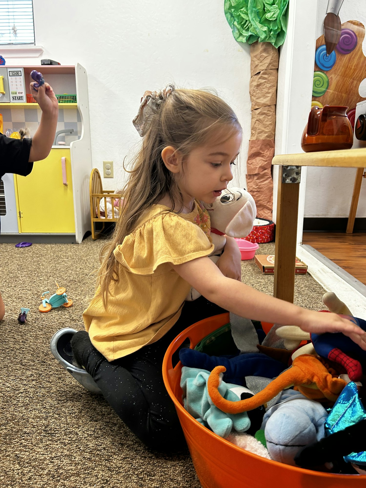 A child in a yellow top is sitting with toys, holding a plush, next to a toy bin in a playroom setting.