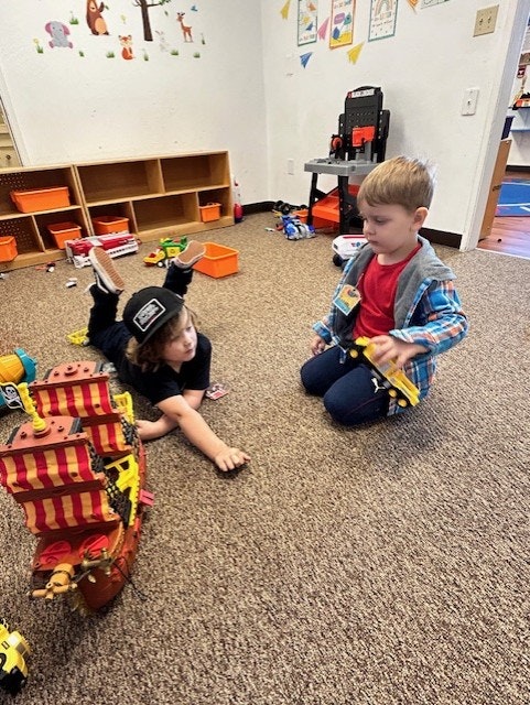 Two kids playing with toys on a carpet in a room with shelves and decorations.
