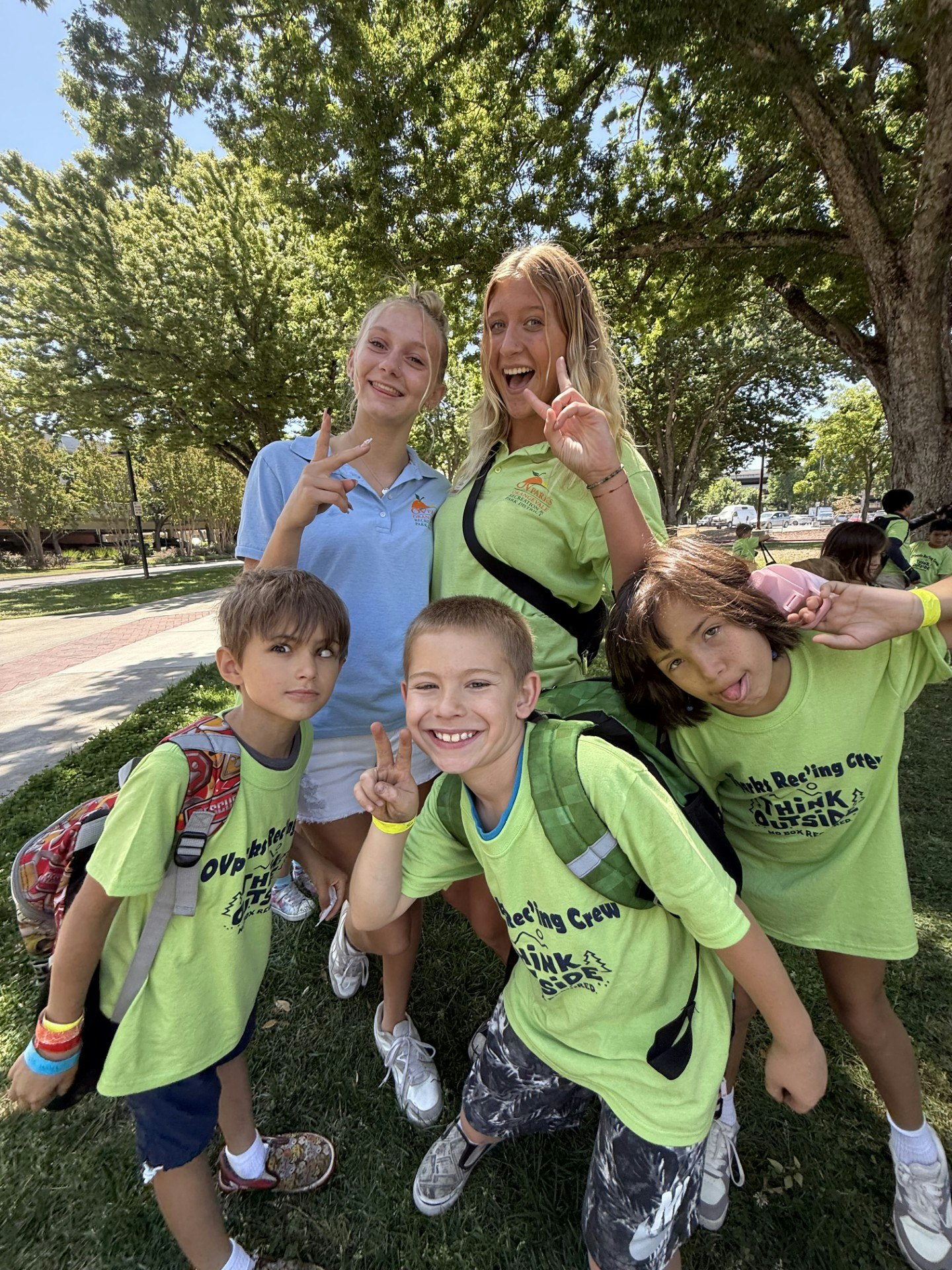 A group of kids and two teens pose playfully outdoors, smiling and showing peace signs, wearing matching bright green shirts.