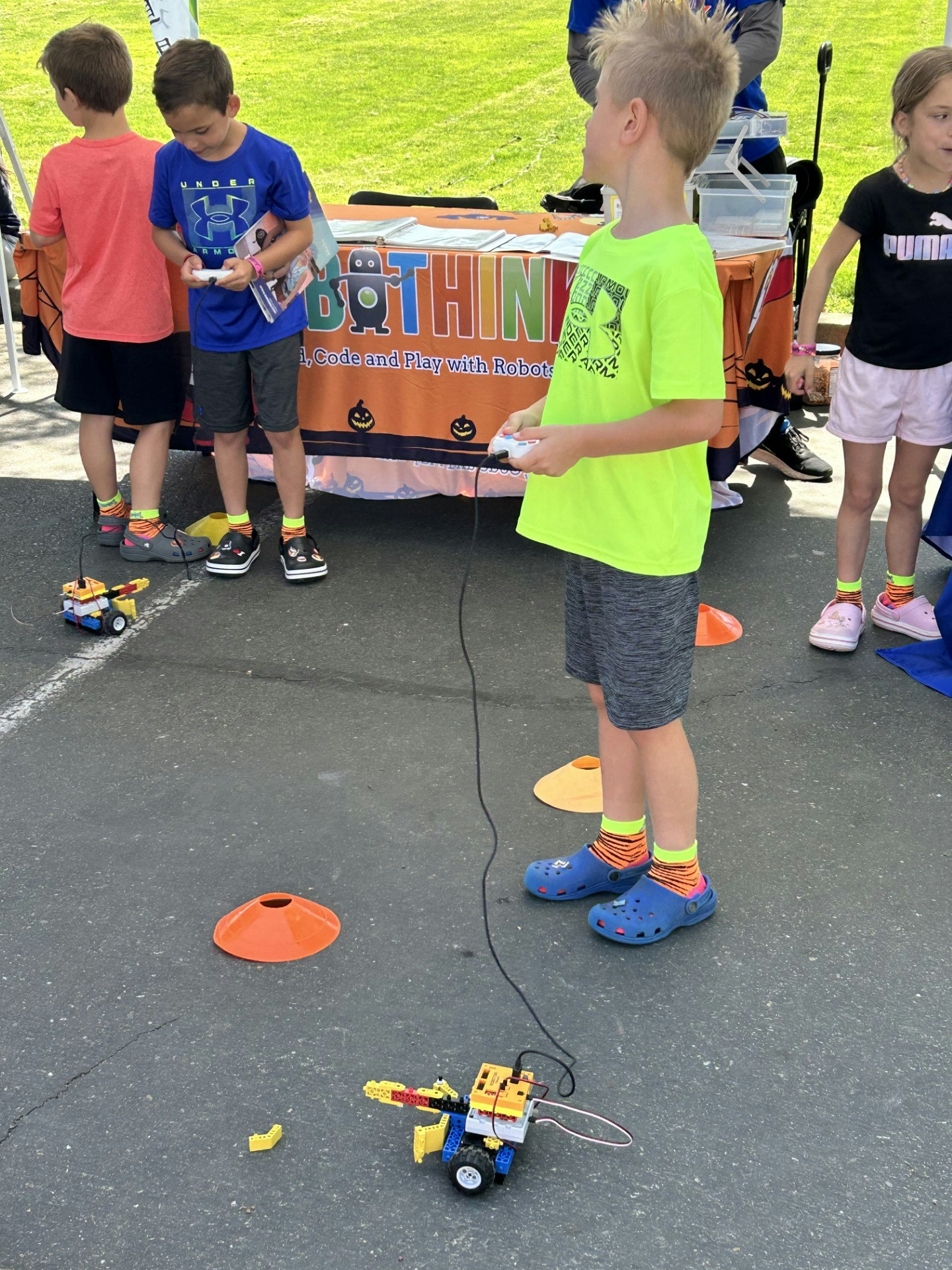 Children controlling small robots with remote controls at an outdoor event.