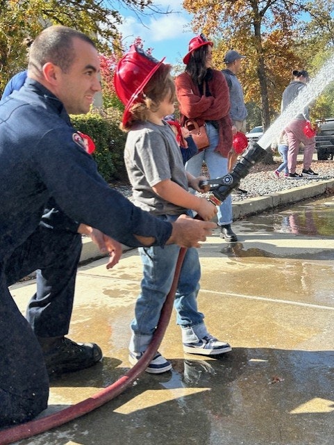 A child with a firefighter helps spray water from a fire hose, wearing a red fire helmet.