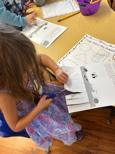 Children cutting and tracing shapes on paper at a classroom table.