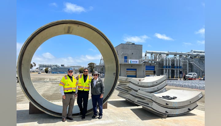 Three people in safety vests posing in front of a large industrial pipe section with equipment in the background.