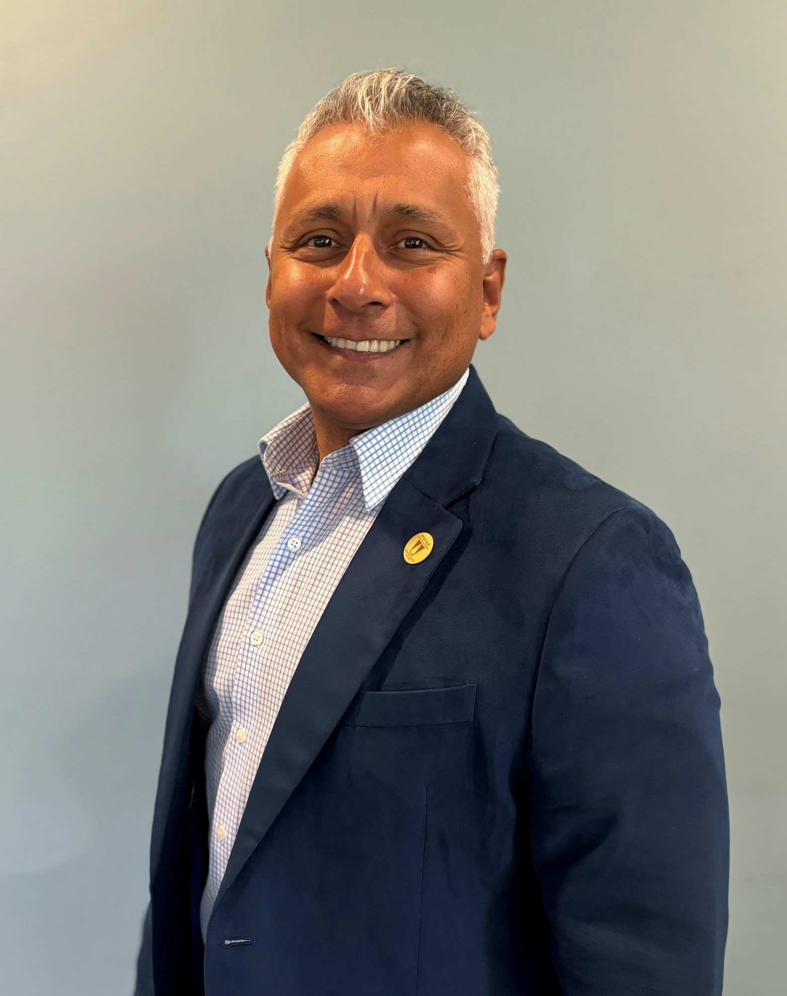 A smiling man in a suit with a pin on his lapel standing against a plain background.