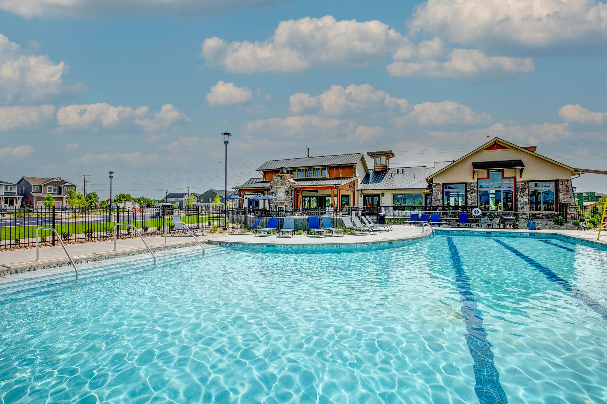 Outdoor pool with clear water, surrounded by lounge chairs, fencing, and buildings in the background under a partly cloudy sky.