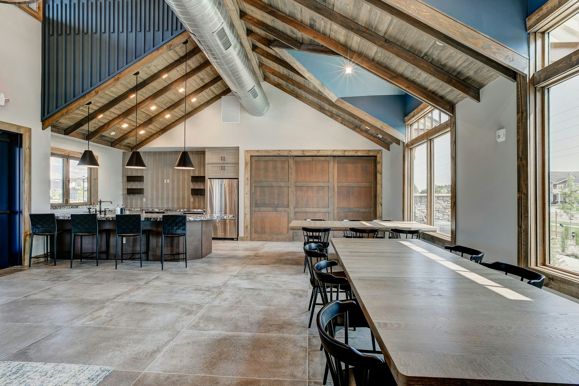 Spacious modern room with wooden beams, a kitchen area, bar stools, long tables, and large windows.