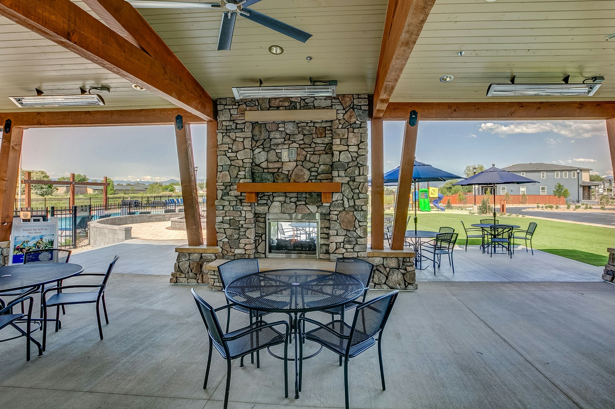 Outdoor pavilion with stone fireplace, round tables, chairs, a playground, and blue umbrellas visible outside.