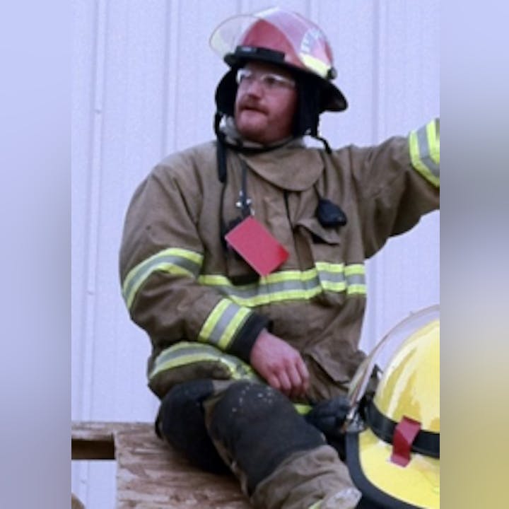 A firefighter in protective gear with a helmet is sitting and looking away.