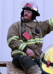 A firefighter in protective gear with a helmet is sitting and looking away.