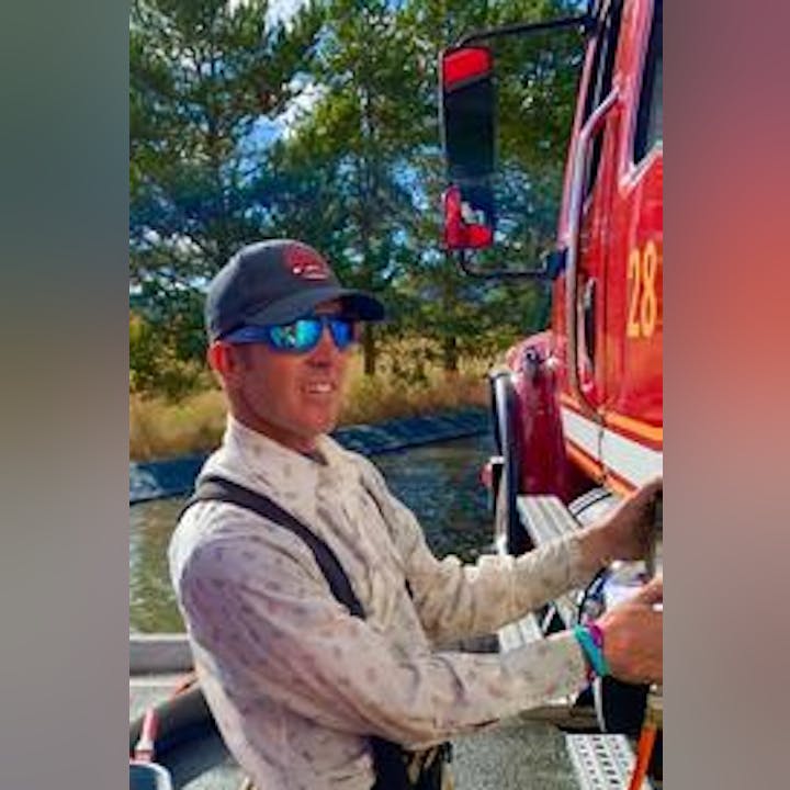A person standing next to a fire truck, wearing sunglasses and a baseball cap, outdoors with trees in the background.