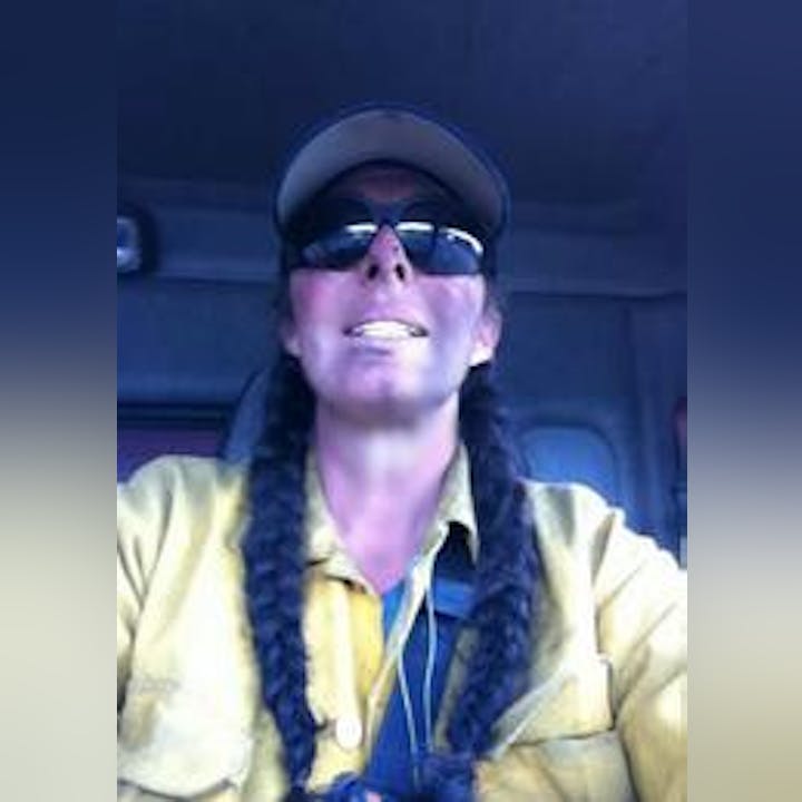 Person in sunglasses and cap, with braids, smiling in a vehicle.