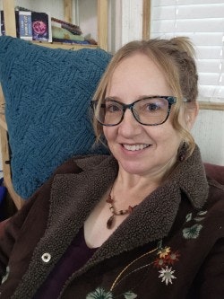 A person wearing glasses and a patterned jacket is smiling, sitting indoors against a blue cushion.