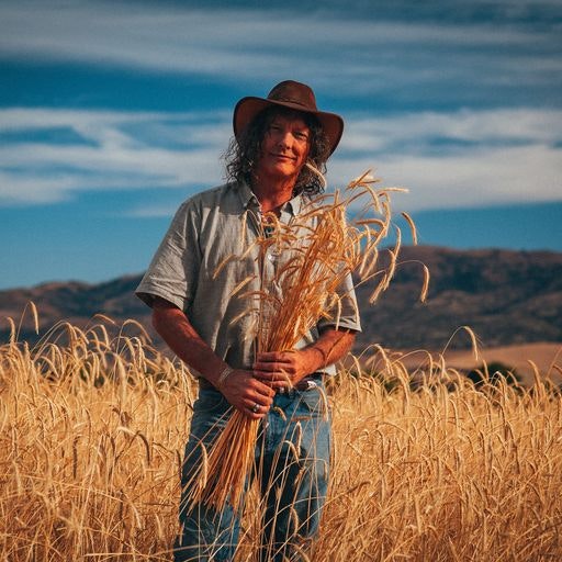 A person stands in a wheat field, holding stalks of wheat, wearing a hat and casual clothes, with a scenic landscape in the background.