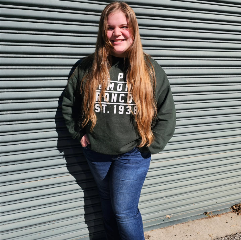A person smiling, leaning against a corrugated metal wall, wearing a dark sweatshirt and jeans.