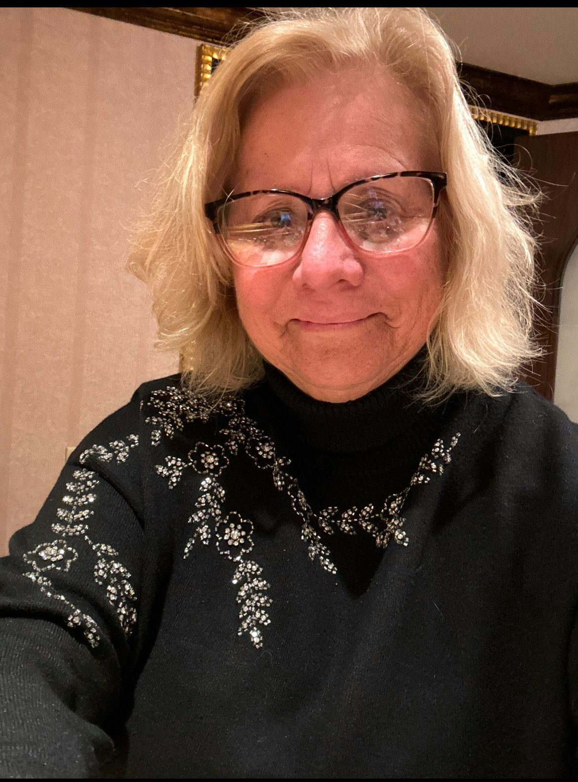 A person with glasses wearing a black sweater with white floral embroidery, smiling at the camera.