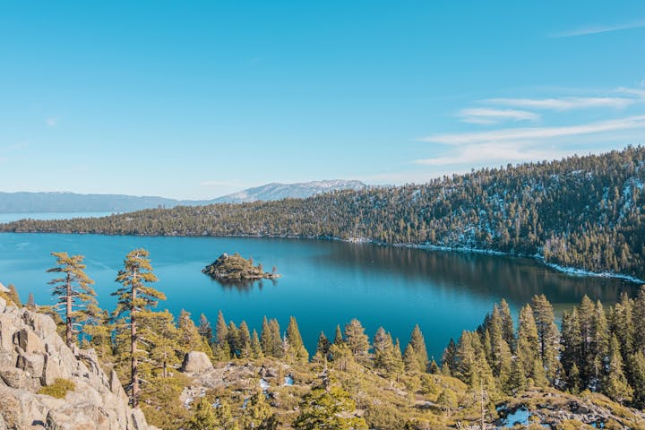 A serene lake with a small island surrounded by pine trees and mountains under a clear blue sky.