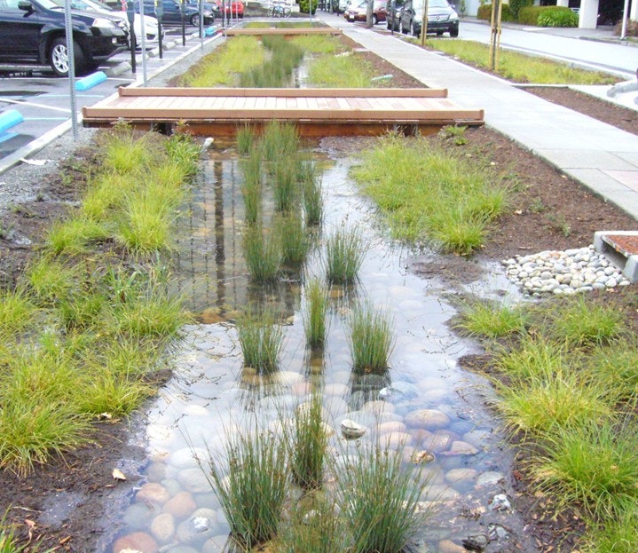 A landscaped drainage ditch with water, rocks, grasses, a wooden footbridge, and nearby parked cars.