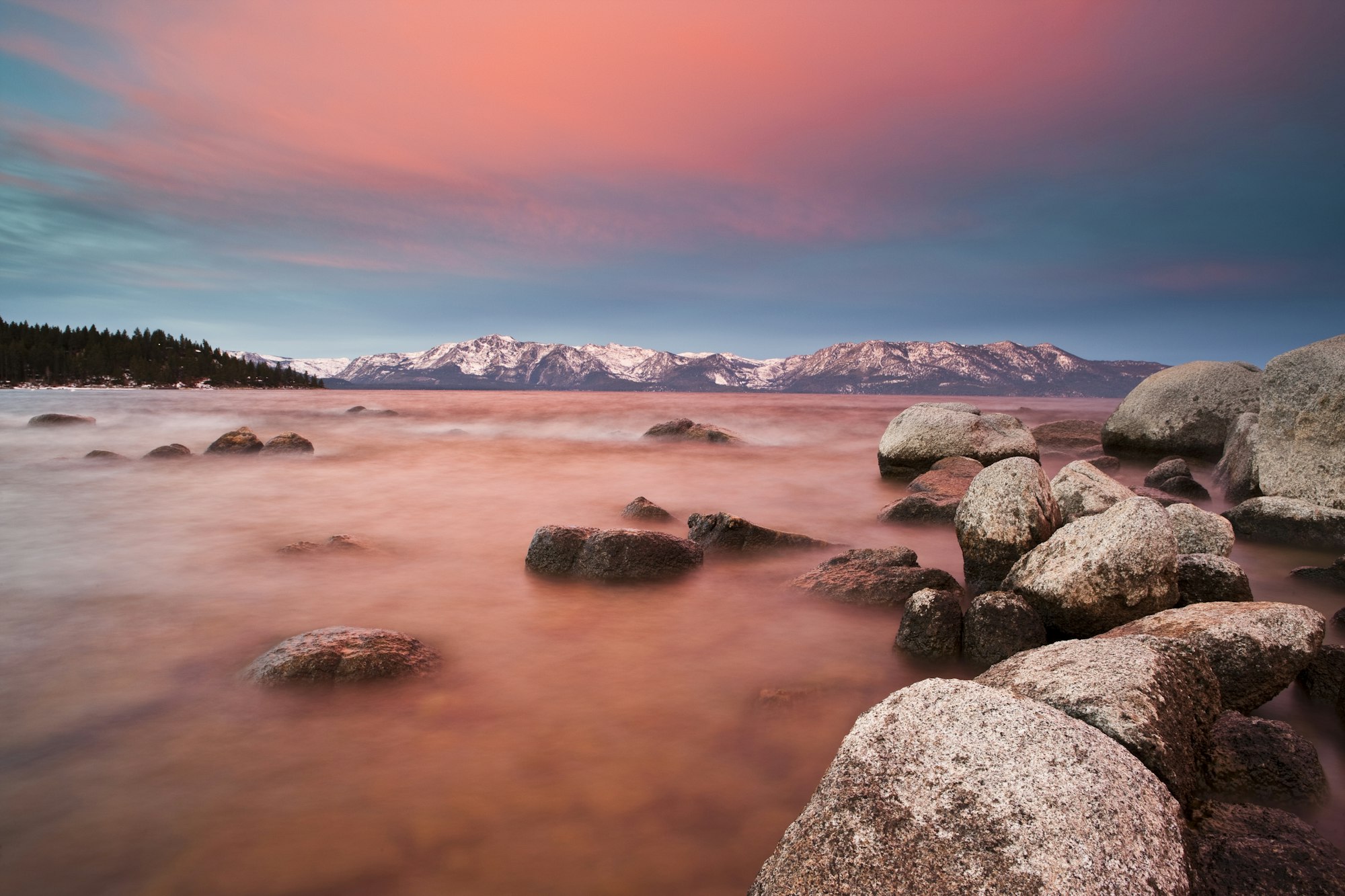 Rocky shoreline, calm water, distant snowy mountains, and a vibrant sunset sky.
