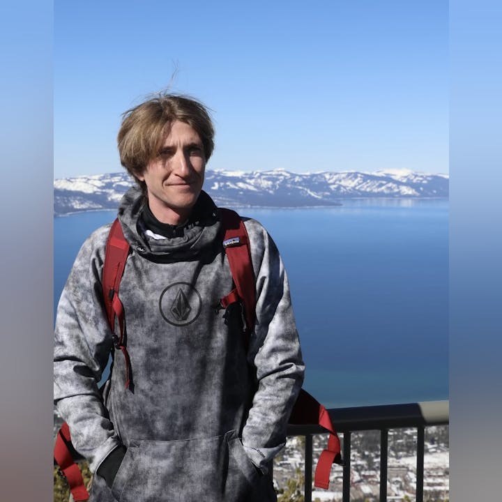 Person in a hoodie with a scenic lake and snow-capped mountains in the background.
