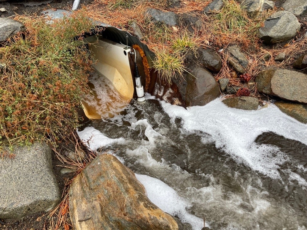 Water flows out of a pipe into a rocky stream with frothy bubbles and surrounding dry plants.