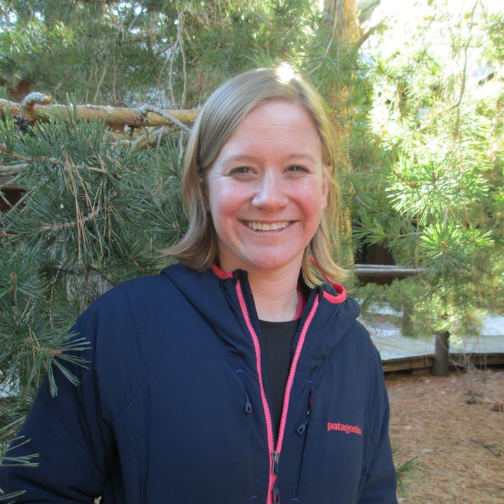 A smiling woman in a blue jacket standing in front of pine trees.