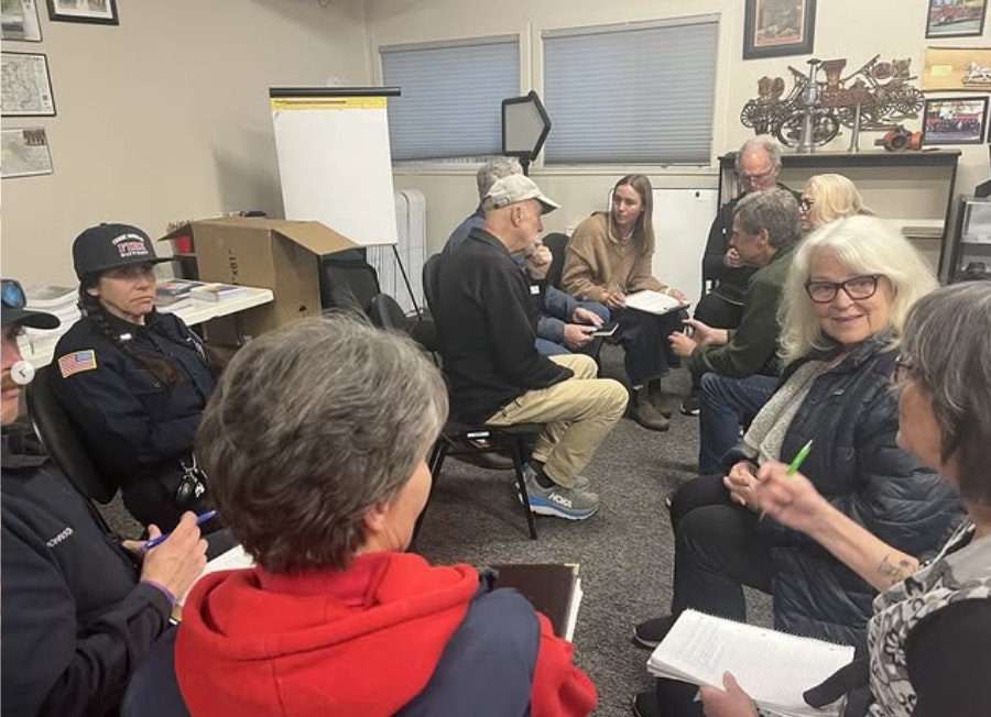 A group discussion is taking place in a room with participants seated, some taking notes, and a person in uniform present.