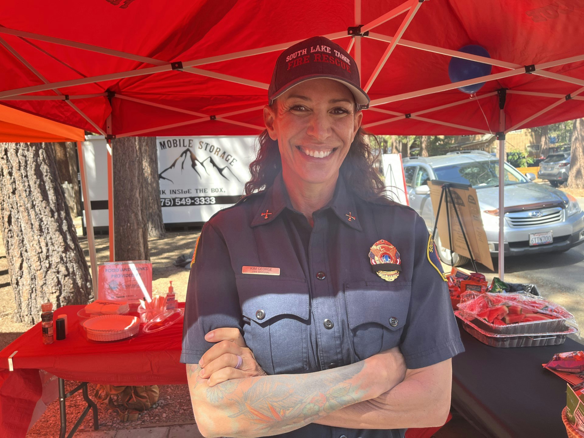 A firefighter smiling under a red tent, wearing a uniform with badges, with a table of supplies and cars in the background.
