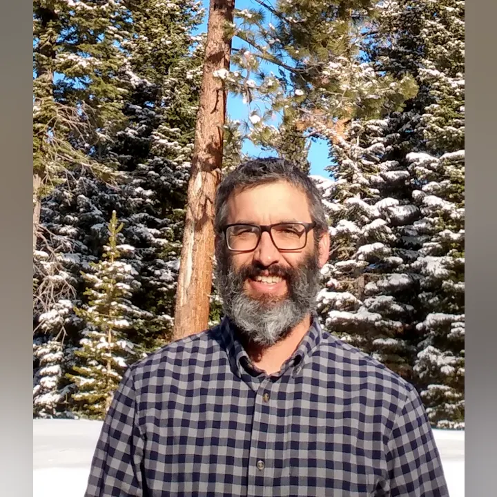 A smiling bearded person in a checkered shirt with snow-covered trees in the background.