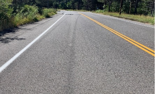 A straight road with yellow and white lines, surrounded by greenery under a clear sky.