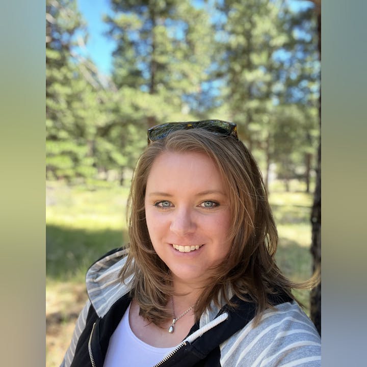 A person smiling in a wooded area with sunglasses on their head.