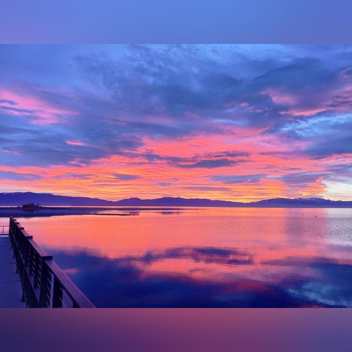 A vibrant sunset with purple and orange hues reflecting on a calm lake with mountains in the background and a railing to the left.