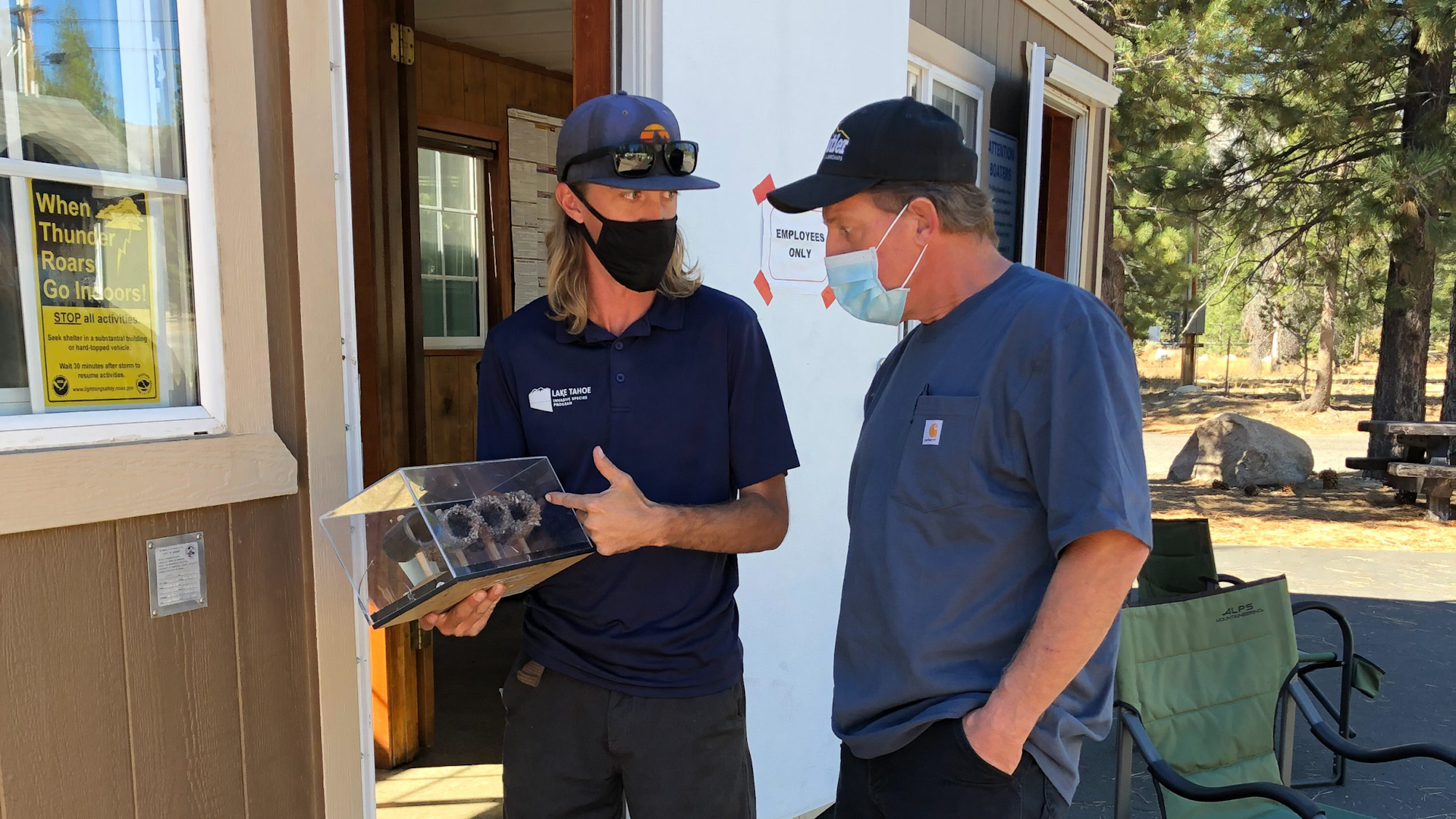 Two men in work attire, one holding a clear box with an item, conversing outside a building.