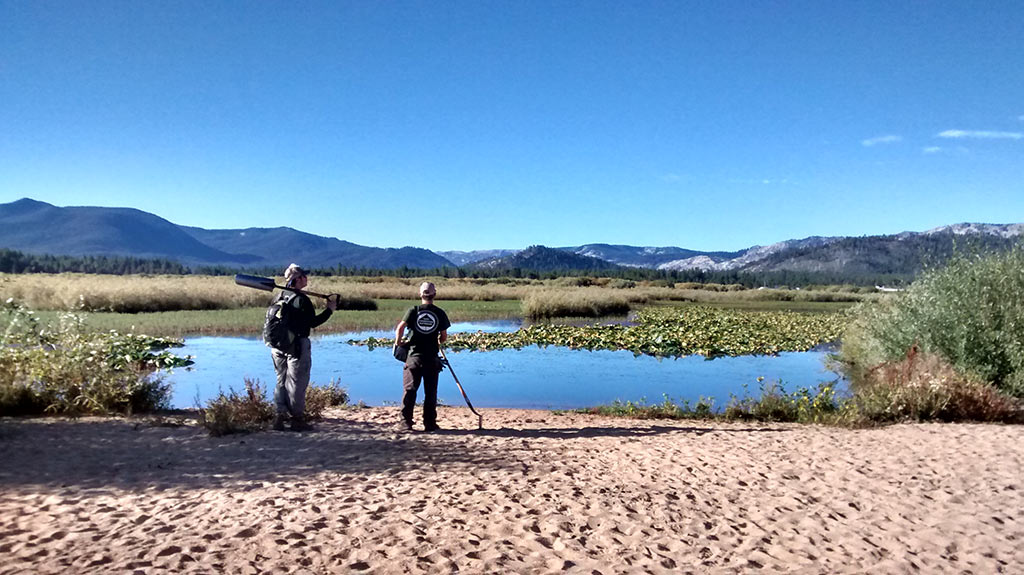 Two people with a canoe by a scenic lake with lily pads, surrounded by mountains and clear blue sky.