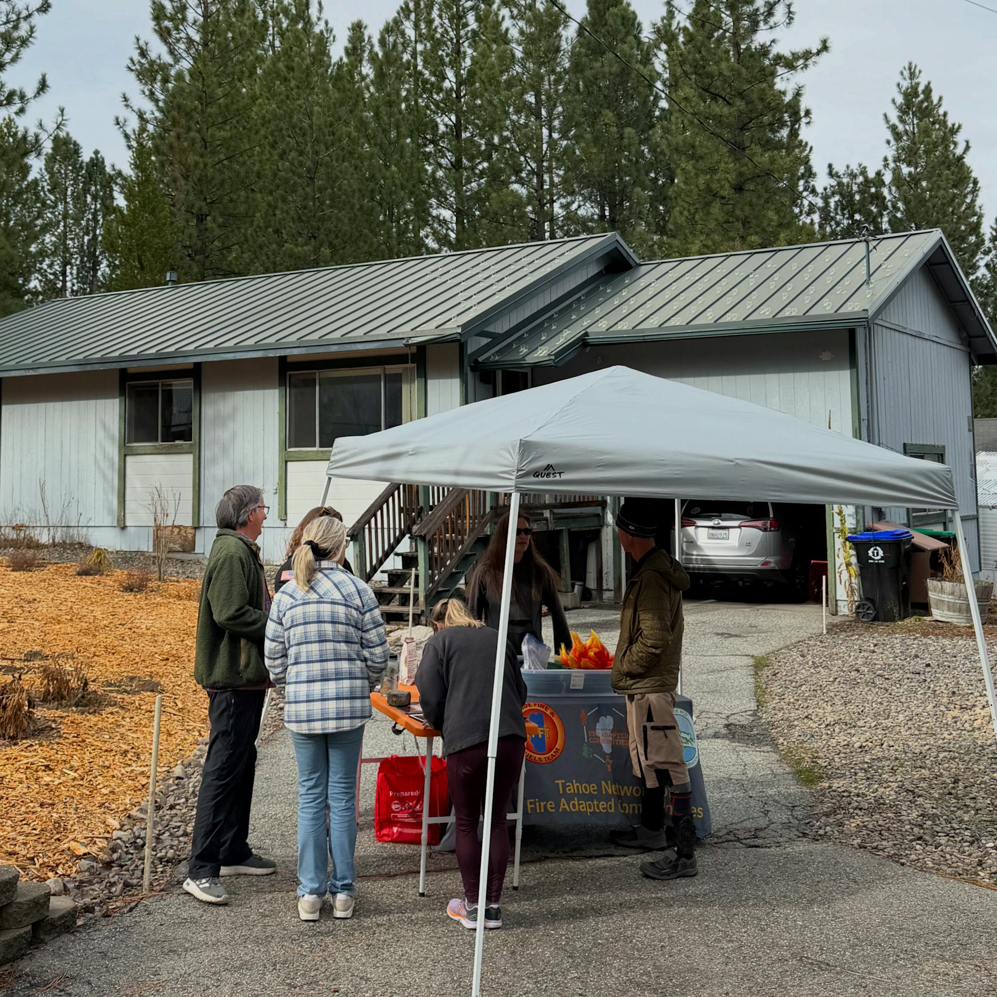 A small gathering under a tent in front of a house, with people interacting and items on a table. Pine trees in the background.