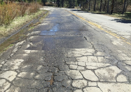 A deteriorated and cracked asphalt road with puddles and a grassy edge.