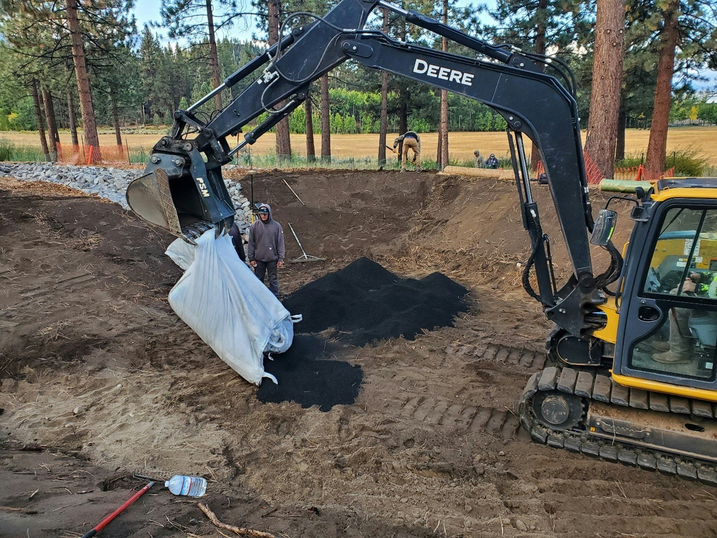 A construction site with a machine lifting a bag of material, surrounded by trees and workers preparing the area.