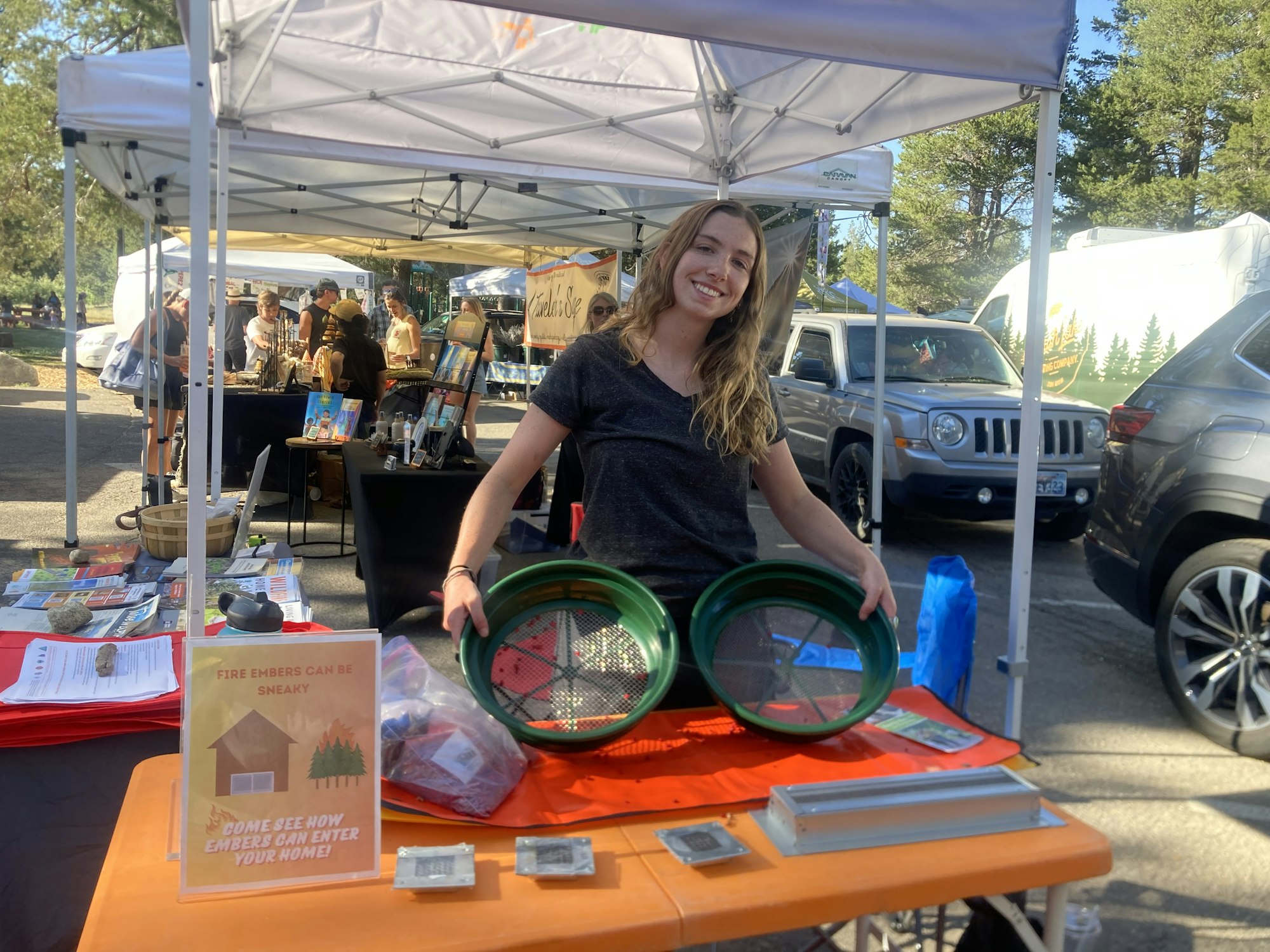 A woman at an outdoor booth holds green mesh trays with informational materials about fire embers and home safety.