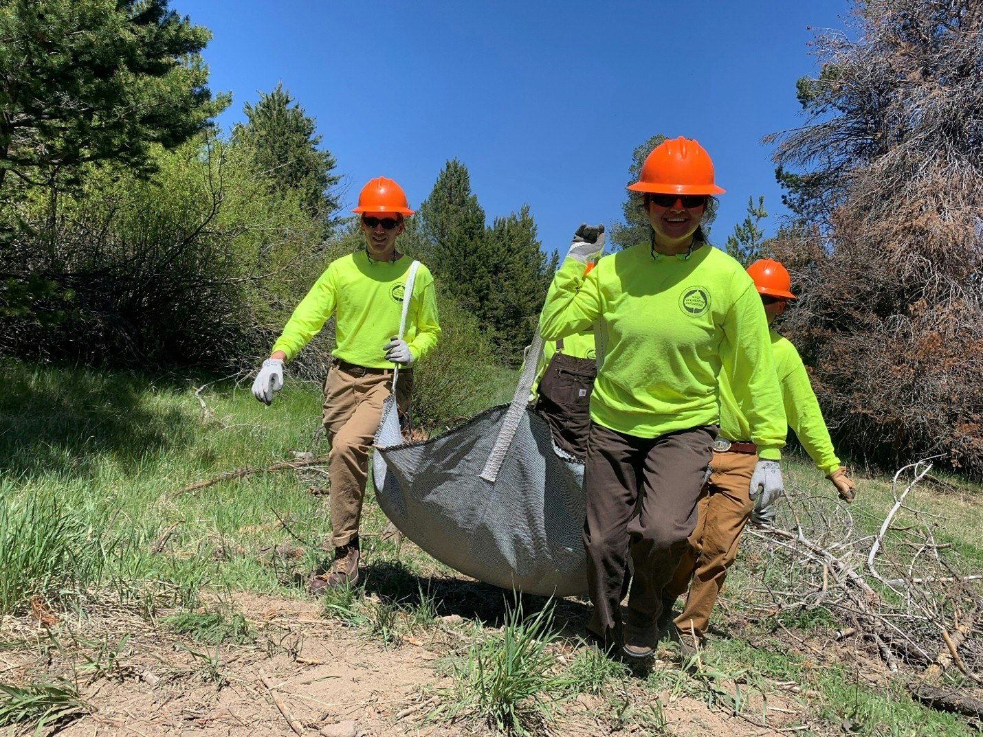 People in bright shirts and hard hats carrying a tarp outdoors in a forested area.