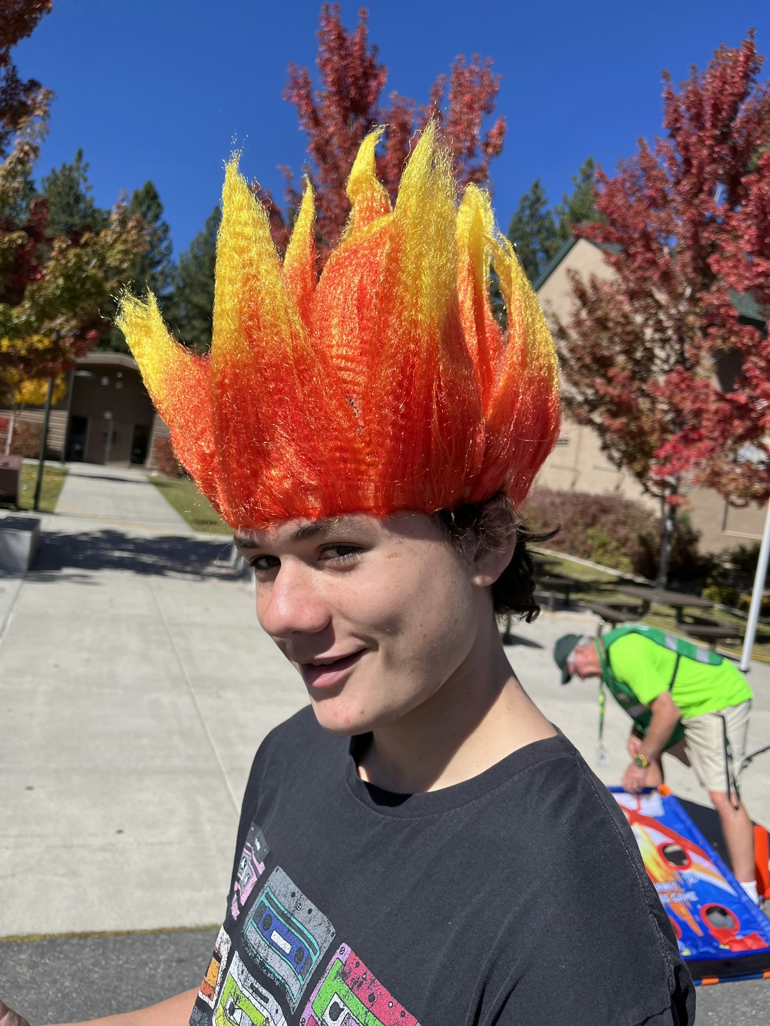 A person wearing a flame-shaped wig outside on a sunny day, with autumn trees and another person in the background.